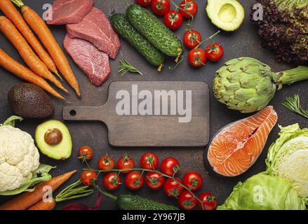 Assortiment de produits crus alimentaires : légumes, viande de bœuf, saumon de poisson et planche à découper en bois vide au centre, vue de dessus de table en pierre brun rustique foncé. Healt Banque D'Images