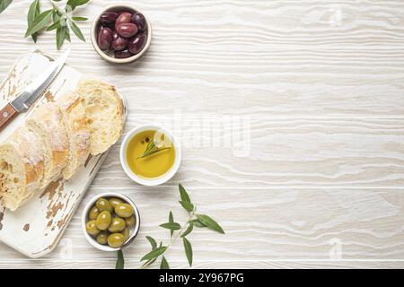 Ciabatta fraîche tranchée sur planche de cuisine à découper, olives vertes et brunes, huile d'olive au romarin, branches d'olivier, fond rustique en bois blanc t Banque D'Images