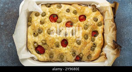 Tête de pain plat traditionnel italien fait maison Focaccia avec olives vertes, huile d'olive, tomates cerises et romarin dans la plaque de cuisson sur rustique sombre Banque D'Images