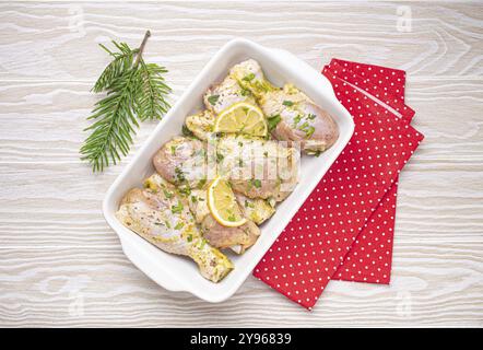 Préparation du repas festif de la veille de Noël avec des pilons de poulet marinés. Cuisses de poulet crues aux herbes, citron dans une casserole en céramique blanche vue de dessus sur Ligh Banque D'Images