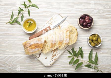 Ciabatta fraîche tranchée sur planche de cuisine à découper, olives vertes et brunes, huile d'olive au romarin, branches d'olivier, fond rustique en bois blanc t Banque D'Images