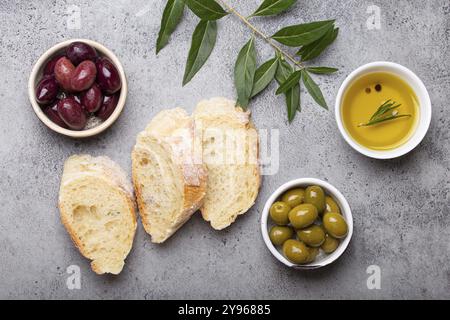 Tranches de ciabatta fraîche, olives vertes et brunes, huile d'olive au romarin, branches d'olivier sur fond rustique de pierre de béton gris au-dessus de la tête, nourriture Banque D'Images