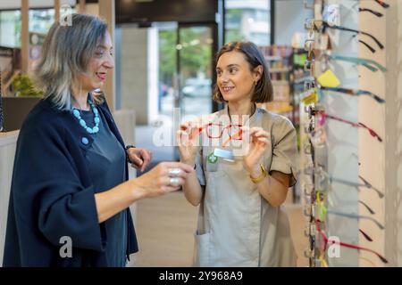 Jeune vendeuse amicale montrant de nouvelles lunettes à un client féminin mature dans le magasin d'optique Banque D'Images