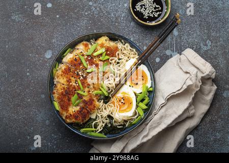 Soupe ramen aux nouilles asiatiques avec filet de poulet panko frit et œufs durs dans un bol en céramique avec des bâtons de côtelette et sauce soja sur fond rustique en pierre Banque D'Images