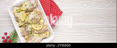 Préparation du repas festif de la veille de Noël avec des pilons de poulet marinés. Cuisses de poulet crues aux herbes, citron dans une casserole blanche vue de dessus sur kitch en bois Banque D'Images