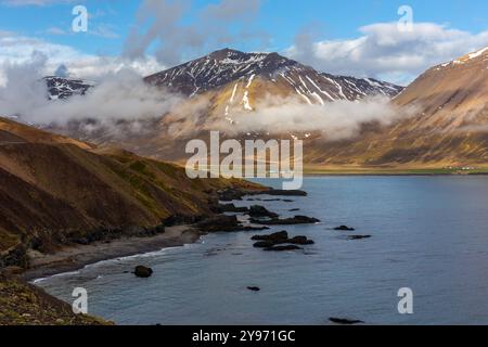 Islande Eastfjords paysage côtier avec montagnes herbeuses couvertes de neige et de nuages et baie d'eau bleue, vue le long de la route 94. Banque D'Images