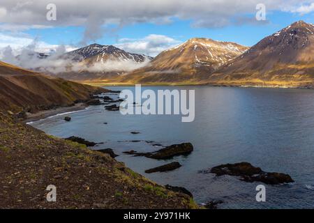 Islande Eastfjords paysage côtier avec montagnes herbeuses couvertes de neige et de nuages et baie d'eau bleue, vue le long de la route 94. Banque D'Images