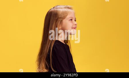 Photo vue de côté de la petite fille charismatique mord ses lèvres, ajoutant une touche ludique sur fond de studio jaune. Banque D'Images