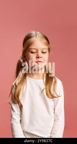 Petite fille avec les cheveux blonds en queue de cochon et chemise blanche regarde vers le bas réfléchi sur fond rose doux, avec une légère expression pensive sur elle Banque D'Images