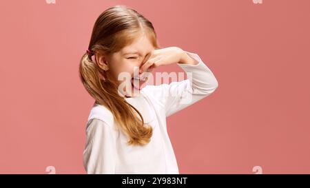 Fille se pince le nez, sort sa langue et plisse les yeux, exprimant le dégoût ou la réaction à la mauvaise odeur sur fond rose pastel, faisant Banque D'Images