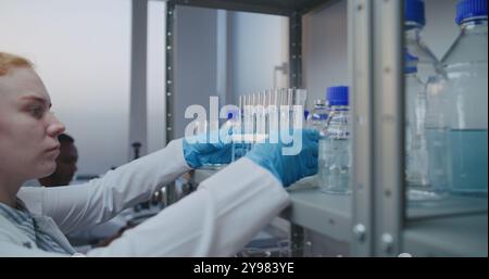 Laboratoire de sciences médicales : chimiste féminine en manteau blanc prend des tubes à essai pour la recherche scientifique de l'étagère avec un équipement de laboratoire. Scientifique afro-américain travaille sur ordinateur en arrière-plan. Banque D'Images