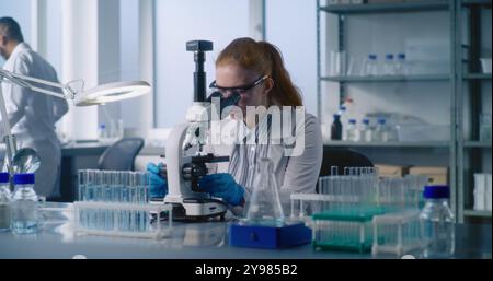 Laboratoire de sciences médicales : microbiologiste féminine prend le liquide du tube à essai, regarde au microscope, analyse l'échantillon à l'aide d'un équipement de haute technologie. Scientifique afro-américain travaille en arrière-plan. Banque D'Images