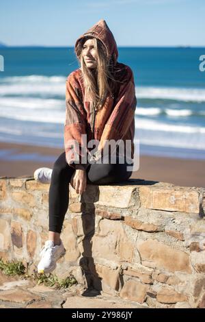 Femme dans une veste à capuche assise sur un mur de pierre près de la plage, profitant d'une journée ensoleillée au bord de l'océan Banque D'Images