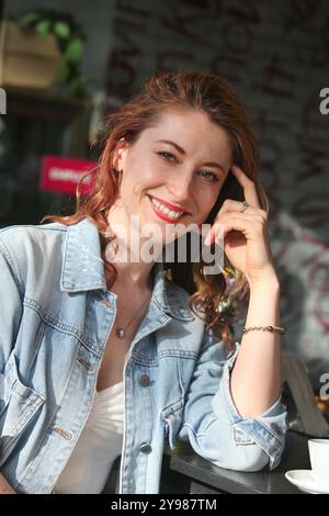 portrait de jolie femme adulte moyenne joyeuse avec des cheveux de gingembre assis à la table dans la cafétéria avec tasse de café à l'extérieur Banque D'Images