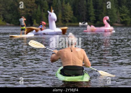 Homme senior pagaie un kayak vert sur un lac, profitant de sa retraite avec des personnes plus jeunes jouant sur des jouets gonflables en arrière-plan. Mise au point sélective Banque D'Images