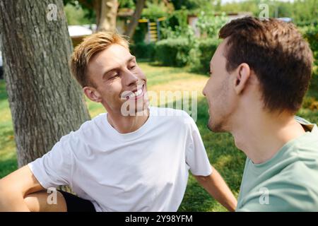 Un joyeux couple gay est assis ensemble dans un parc luxuriant, partageant sourires et chaleur dans la lumière du soleil. Banque D'Images