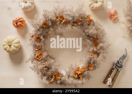 Couronne avec des fleurs oranges et des matériaux naturels secs sur fond beige en bois. Vue de dessus. BRICOLAGE. Atelier pour la décoration de jour de Thanksgiving faite à la main. Banque D'Images