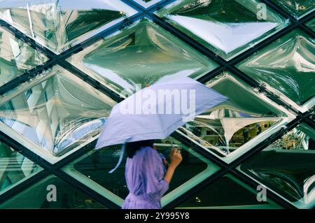 Une jeune fille tenant un parapluie marche contre une façade hexagonale en verre, capturant un moment d'élégance au milieu de l'architecture urbaine Banque D'Images