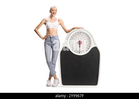 Femme mince heureuse en jeans et top crop debout à côté d'une balance de poids isolé sur fond blanc Banque D'Images