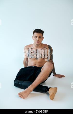 Un jeune homme avec des tatouages complexes présente sa jambe prothétique assis dans un studio lumineux. Banque D'Images