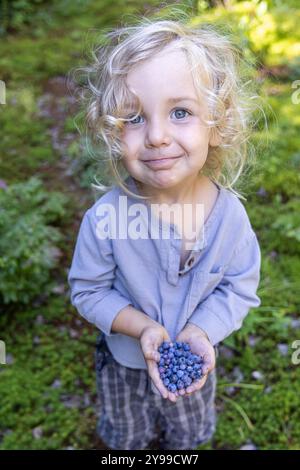 Le jeune garçon tient une poignée de bleuets fraîchement cueillis tout en se tenant debout dans une forêt. Mise au point sélective Banque D'Images