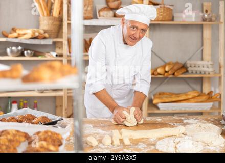 Homme senior travaille dans la boulangerie comme boulanger, pétrit la pâte, travaille avec la farine. Banque D'Images