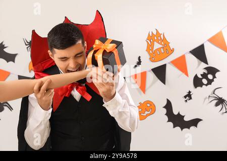 Jeune homme habillé pour Halloween comme vampire mordant la main avec cadeau sur fond clair Banque D'Images