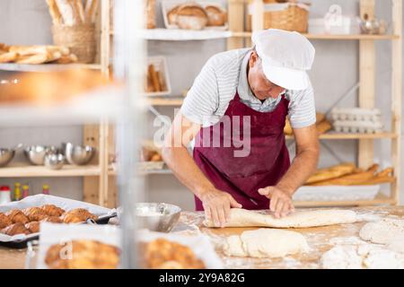 Homme senior travaille comme boulanger dans la boulangerie, moule les tartes à partir de pâte, crée des croissants à partir de pâte. Banque D'Images