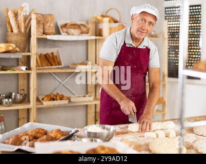 Homme senior travaille dans la boulangerie comme boulanger, coupe la pâte en portions, forme des morceaux de pâte pour créer des petits pains. Banque D'Images