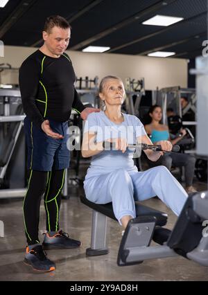 Entraîneur de fitness masculin est engagé explique comment exercer sur le simulateur à une femme âgée dans le gymnase Banque D'Images