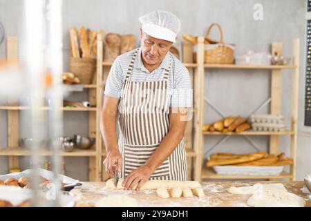 Homme senior travaille dans la boulangerie comme boulanger, coupe la pâte en portions, forme des morceaux de pâte pour créer des petits pains. Banque D'Images