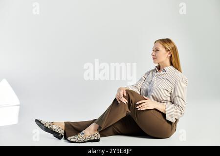 Une femme élégante aux cheveux longs s'assoit gracieusement dans un studio gris, portant une chemise rayée et un pantalon. Banque D'Images