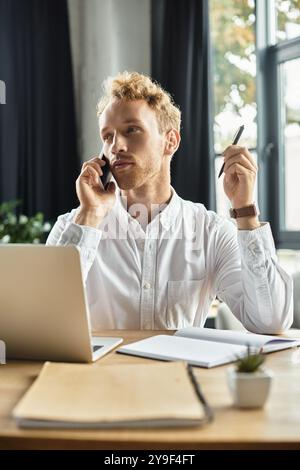 Une rousse réfléchie dans une chemise blanche s'engage dans un projet de travail tout en faisant un appel téléphonique dans un bureau. Banque D'Images