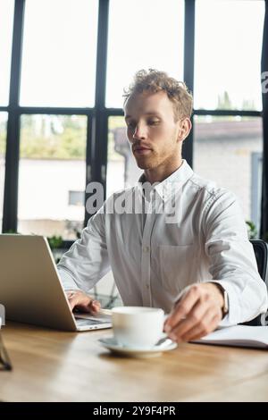 Une rousse dans une chemise blanche se concentre sur son ordinateur portable tout en dégustant un café dans un espace de travail contemporain. Banque D'Images