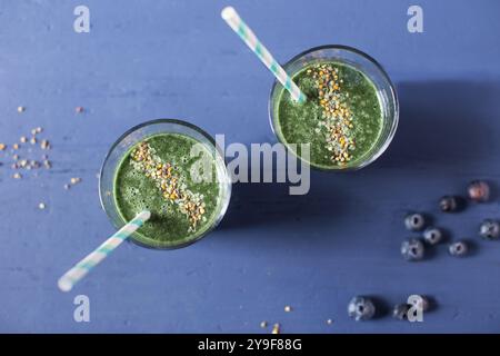 Deux verres avec un smoothie vert surmonté de pollen d'abeille et de graines de chanvre avec des pailles rayées vertes et blanches tirées au-dessus sur fond bleu. Banque D'Images