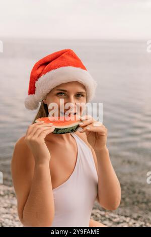 Une femme mange une pastèque tout en portant un chapeau de père noël rouge. La scène est légère et festive, car la femme profite d'une collation rafraîchissante sur un Banque D'Images