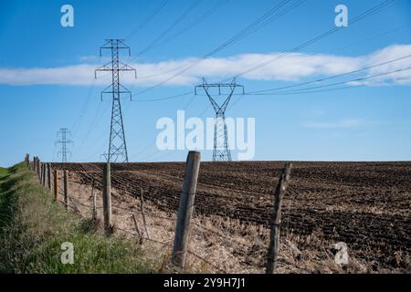 Des lignes électriques bourdonnantes maintenues en hauteur par de hautes tours électriques en treillis d'acier sur un champ labouré avec une clôture en fil de fer barbelé dans les prairies canadiennes à Rocky View Count Banque D'Images