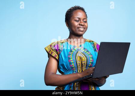 Jeune adulte en vêtements ethniques afro-américains tient un ordinateur portable en studio, en utilisant le réseau Internet pour vérifier les médias sociaux et les sites Web. Femme moderne souriante et profitant de la nouvelle technologie sans fil. Banque D'Images