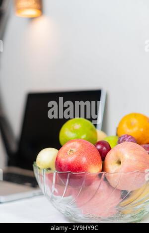 Un bol en verre transparent rempli de divers fruits, y compris des pommes rouges, des pommes vertes, des raisins et une orange, placé sur une table près d'un ordinateur portable. Le backgrou Banque D'Images