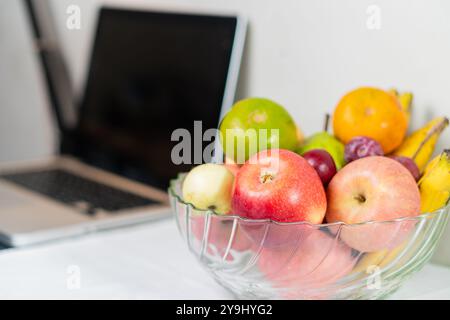 Un bol en verre transparent rempli d'une variété de fruits frais, y compris des pommes rouges et vertes, des bananes, des raisins et une orange, placé à côté d'un ordinateur portable sur un Banque D'Images