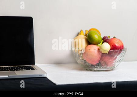 Un bol en verre transparent rempli de divers fruits, y compris des pommes, des bananes et des agrumes, placé à côté d'un ordinateur portable sur une nappe blanche. Banque D'Images