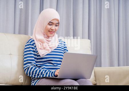 Femme musulmane asiatique portant le hijab assis à l'aide de l'ordinateur portable pour faire le travail, travaillant de la maison à sa maison sur le canapé et s'asseoir et regarder la télévision. Banque D'Images