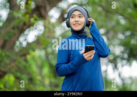 Femme musulmane mignonne asiatique portant des vêtements de sport et un hijab bleu s'entraînant et portant des écouteurs écoutant de la musique sur son téléphone à l'extérieur dans le parc Banque D'Images