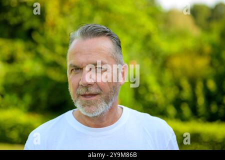 Un homme mature avec des cheveux gris et une barbe, portant un T-shirt blanc, se tient à l'extérieur avec un fond vert éclatant et flou. Banque D'Images