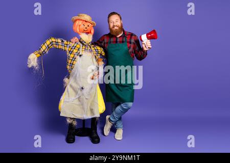 Photo de marron barbu jardinier homme câlin avec épouvantail saisonnier offre d'halloween événement isolé sur fond de couleur pourpre Banque D'Images