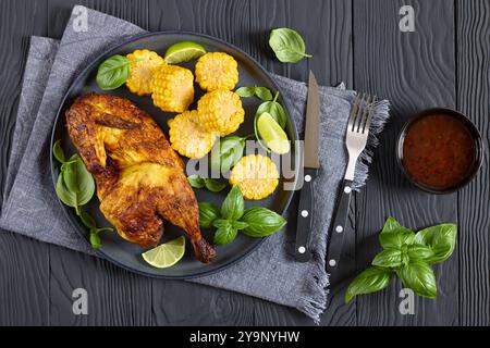 la moitié de poulet rôti avec des pièces d'épis de maïs et des feuilles de basilic frais sur une assiette sur une table en bois noir avec couverts et sauce chili douce, vue horizontale Banque D'Images