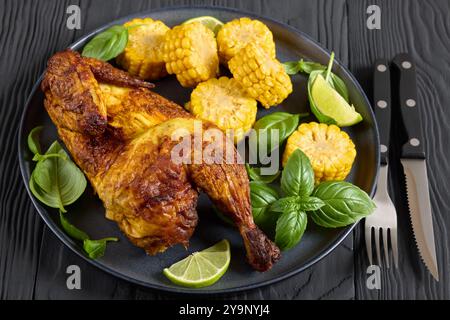 la moitié du poulet rôti avec des pièces d'épis de maïs et des feuilles de basilic frais sur l'assiette sur une table en bois noir avec des couverts, gros plan Banque D'Images
