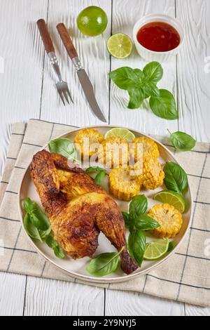 la moitié de poulet rôti avec des pièces d'épis de maïs et des feuilles de basilic frais sur une assiette sur une table en bois blanc avec couverts, citron vert et sauce chili doux, verticale Banque D'Images