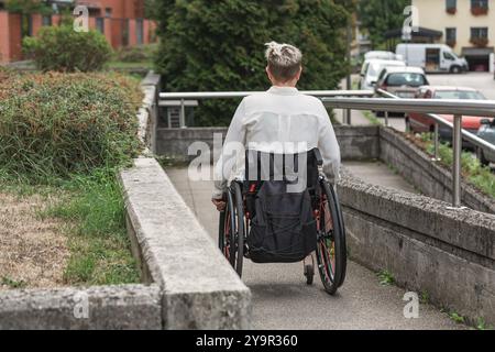 Personne ayant une déficience physique conduisant un fauteuil roulant le long d'une rampe accessible pour accéder à la rue. Handicap et concepts de mobilité. Banque D'Images