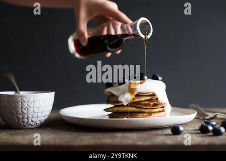 Pile de crêpes à la banane et au sarrasin avec yaourt, myrtilles et sirop d'érable. Main de femme versant du sirop d'érable sur des crêpes. Banque D'Images